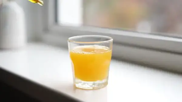 Golden liquid falling from a glass pipette into a small glass of orange juice on a kitchen windowsill